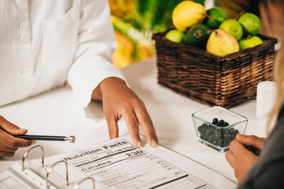 a woman at a table in a white doctor's coat with a patient and a binder there is also fruit on the table 