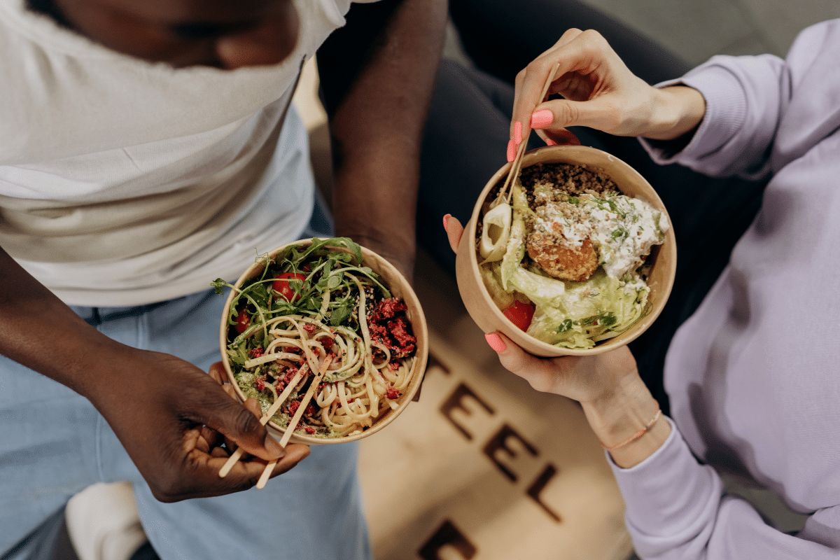 two women with bowls in front of them they are both eating different foods one has pasta the other has salad 